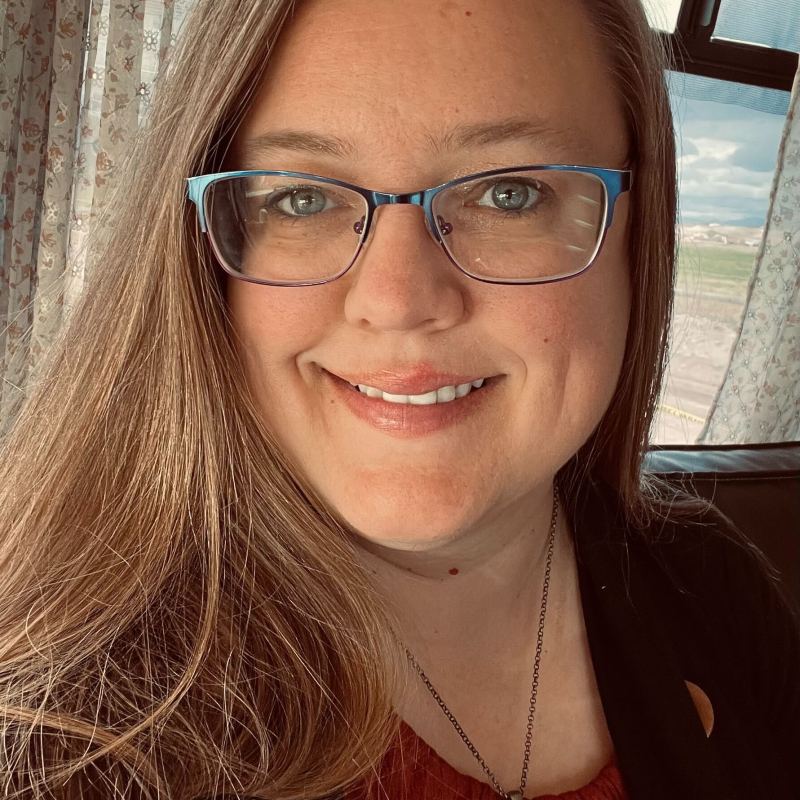 Person with glasses and long hair smiling indoors near a window.