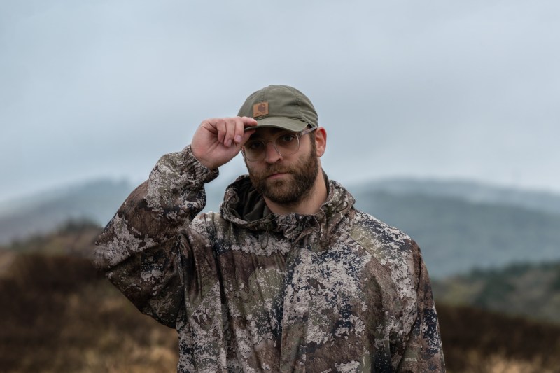 Person in camouflage jacket and cap standing outdoors on a cloudy day.
