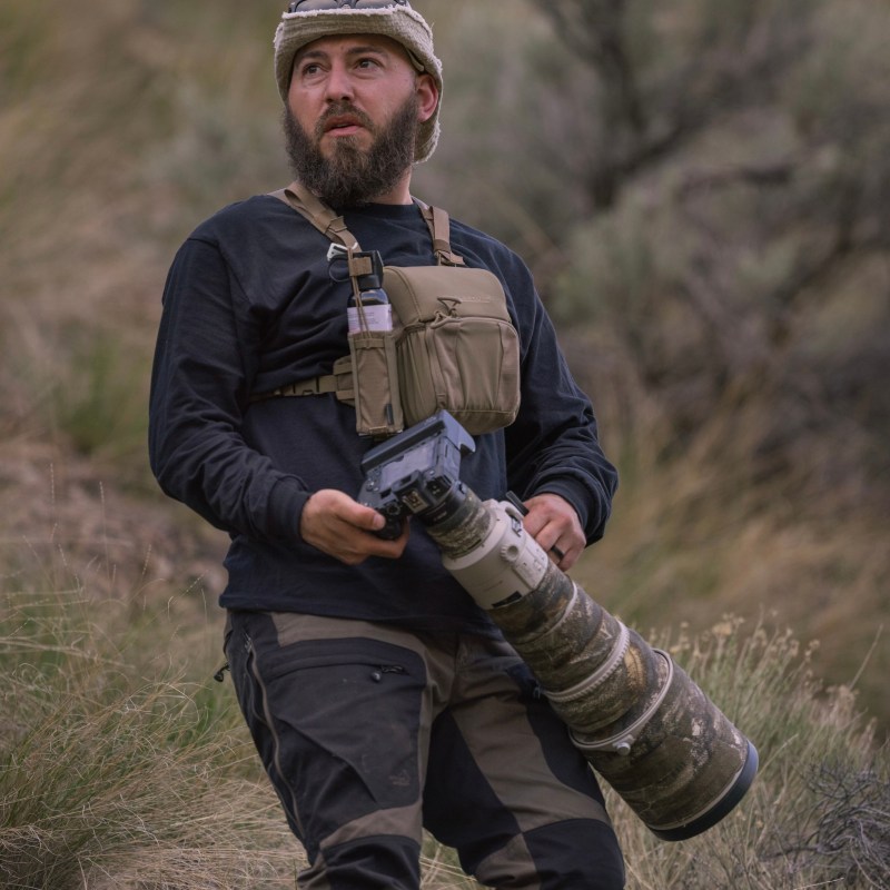 Man outdoors with camouflage gear holding large camera lens.