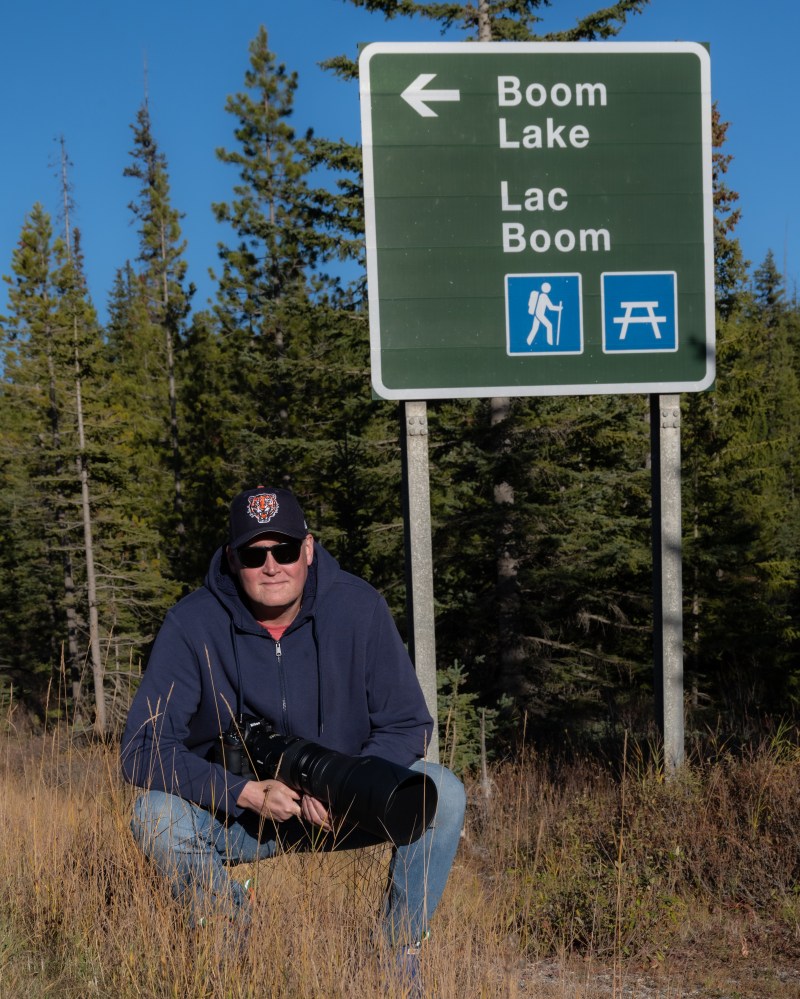 Person crouching near Boom Lake sign with hiking and picnic icons, surrounded by trees.